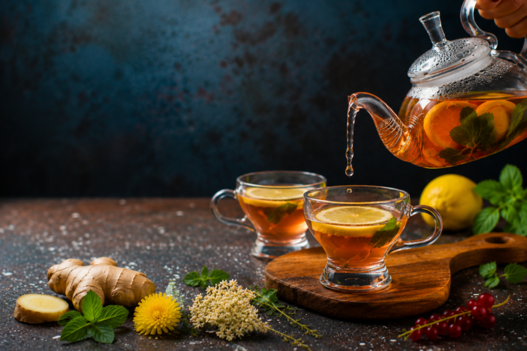 Glass teapot pouring herbal lymphatic drainage tea into a clear cup with lemon, ginger, mint, and fresh herbs on a rustic wooden board.